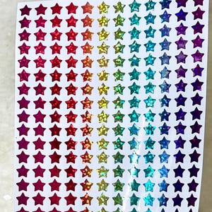 May include: A sheet of rainbow holographic star stickers. The stars are arranged in rows and columns, with each row featuring a different color of the rainbow. The stars are iridescent and sparkle in the light.