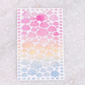 May include: A sheet of cloud-shaped stickers in a gradient of colors. The stickers transition from pink to yellow to blue, with glitter accents. The stickers are arranged in a rectangular pattern.