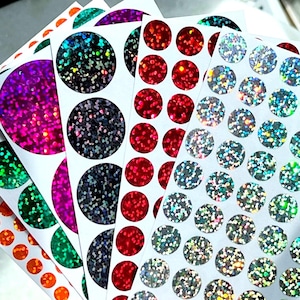 May include: A set of six sheets of iridescent holographic stickers. Each sheet features a different color of round stickers with a glittery finish. The colors are pink, green, blue, red, black, and silver.