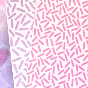 May include: Close-up of two white cards with a repeating pattern of pink and lavender glittery dashes. The dashes are scattered across the cards, creating a textured effect. The background includes a glimpse of a patterned fabric.