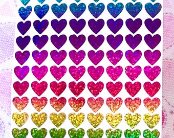 Könnte beinhalten: Eine Bogen mit regenbogenfarbenen, holografischen Herzaufklebern. Die Herzen sind in Reihen angeordnet, wobei jede Reihe eine andere Farbe des Regenbogens zeigt. Die Farben sind Blau, Lila, Rosa, Rot, Orange, Gelb und Grün.