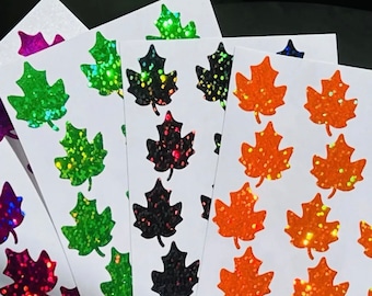 Könnte beinhalten: Fünf Blätter weißen Papiers mit Reihen von schimmernden, holografischen Ahornblattaufklebern. Die Aufkleber sind in fünf Farben: grün, schwarz, orange, lila und blau.