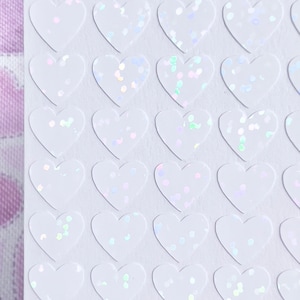 May include: A sheet of white heart-shaped stickers with iridescent glitter. The stickers are arranged in a grid pattern.