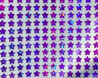 Könnte beinhalten: Ein Bogen mit lila sternförmigen Aufklebern mit glitzerndem, schillerndem Finish. Die Aufkleber sind in einem Rastermuster angeordnet, wodurch ein sich wiederholendes Design entsteht. Die Sterne haben eine glänzende, reflektierende Oberfläche, und der Gesamteffekt ist auffällig und dekorativ.