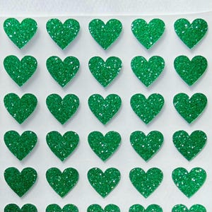 May include: Close-up of a sheet of green glitter heart stickers. The hearts are evenly spaced and have a sparkling texture. The stickers are a vibrant green color, perfect for crafts or decorations.