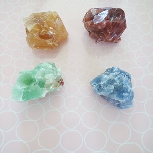 Pode incluir: Quatro cristais de diferentes cores sobre um fundo rosa com um padrão de círculos brancos. Os cristais são de cor castanho claro, vermelho escuro, verde claro e azul claro.