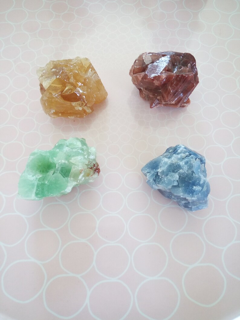 May include: Four different colored crystals on a pink background with a white circle pattern. The crystals are a light brown, a dark red, a light green, and a light blue.