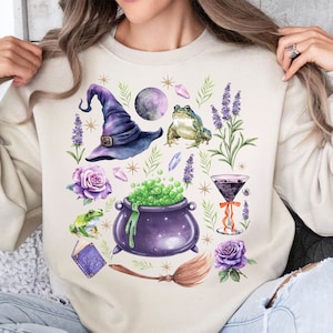 May include: A cream-colored sweatshirt with a whimsical design featuring a witch's hat, a cauldron, a frog, a rose, a moon, and a broom. The design is in shades of purple, green, and brown.