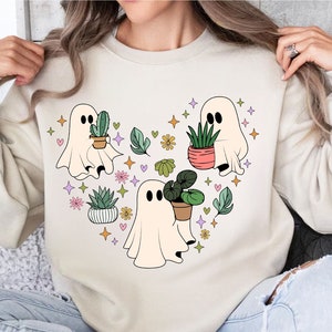 Puede incluir: Una sudadera de color crema con un diseño gráfico de tres fantasmas sosteniendo plantas en macetas. Los fantasmas son blancos y las plantas son verdes, rosas y naranjas. El fondo está lleno de pequeños corazones, estrellas y hojas.