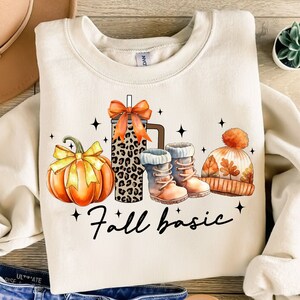 Könnte beinhalten: Ein cremefarbener Pullover mit einem grafischen Design aus einem Kürbis, einer Kaffeetasse mit Leopardenmuster, einem Paar braunen Stiefeln und einer braunen und orangen Mütze. Der Text "fall basic" ist unter dem Grafikdruck aufgedruckt.