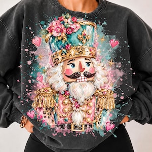 Op de afbeelding: Een antracietgrijs sweatshirt met ronde hals en een levendige aquarel-afbeelding van een notenkraker-soldaat. De notenkraker draagt een blauwgroene hoed versierd met roze bloemen, gouden accenten en luipaardprint, omringd door roze hartjes.