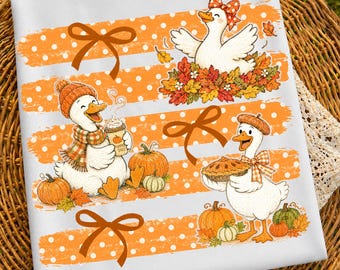 Op de afbeelding: Een wit t-shirt met een herhalend patroon van oranje horizontale strepen met witte stippen en bruine strikken. Het ontwerp bevat drie witte eenden, één met een muts en sjaal met een koffiebeker, één met een strik en één met een taart.