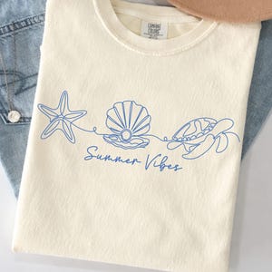 May include: Cream-colored t-shirt with a blue line drawing of a starfish, seashell, and turtle, with the words "Summer Vibes" in script. The shirt is made of a soft material and is perfect for summer.