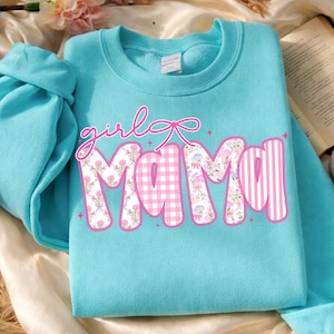 Puede incluir: Una sudadera azul claro con las palabras "girl mama" en una fuente decorativa. La palabra "mama" est&aacute; dise&ntilde;ada con patrones florales, de cuadros y de rayas en rosa y blanco. Una cinta rosa est&aacute; encima de la palabra "mama".