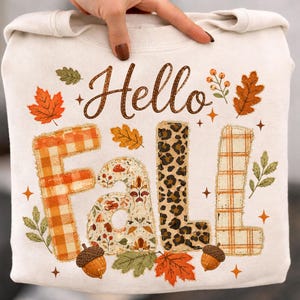 以下が含まれることがあります： 茶色の筆記体でHello Fallと書かれたクリーム色のスウェットシャツ。Fallという単語は、オレンジ色のチェック柄、キノコ柄、ヒョウ柄、ベージュのチェック柄という異なるパターンの4つの大きな文字で構成されており、周囲には秋の葉が描かれています。