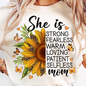 May include: Cream-colored t-shirt featuring a sunflower design with leopard print accents and the words "She is" above a list of positive attributes. Orange butterflies and hearts decorate the design. The word "mom" is at the bottom.
