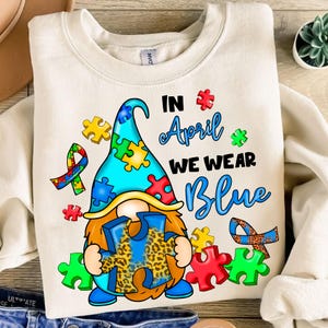 Pu&ograve; includere: Una felpa bianca con un gnomo blu e giallo che tiene un pezzo di puzzle blu con un motivo a stampa leopardata. Il testo "In April We Wear Blue" &egrave; stampato sulla felpa.