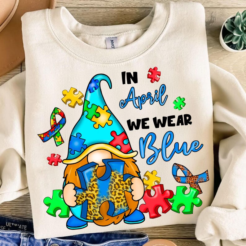 Pu&ograve; includere: Una felpa bianca con un gnomo blu e giallo che tiene un pezzo di puzzle blu con un motivo a stampa leopardata. Il testo "In April We Wear Blue" &egrave; stampato sulla felpa.
