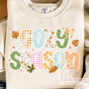 May include: Cream-colored sweatshirt with the words "COZY SEASON" in a patchwork design. The letters are decorated with floral, gingham, and polka dot patterns, with fall leaf and bow accents. The design includes the text "RETROSUBLIMATION DIGITAL DESIGN EST. 2020".