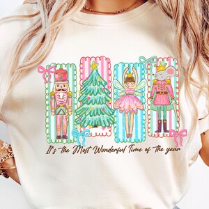 May include: Cream-colored t-shirt featuring a festive design with a nutcracker, Christmas tree, fairy, and mouse in pink and green frames. The text "It's the Most Wonderful Time of the year" is printed below the illustrations.