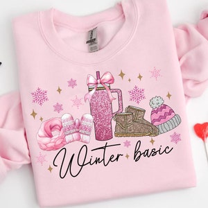 May include: Pink sweatshirt with a winter-themed design. The design features a pink glitter tumbler, a pair of brown boots, pink mittens, a pink scarf, and a pink and white hat. The text "Winter basic" is written in black cursive lettering.