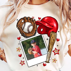 May include: Cream-colored t-shirt featuring a baseball-themed graphic. The design includes a baseball, glove, red helmet, and a wooden bat. A photo of a child in a baseball uniform is also included. The design is decorated with stars and hearts.
