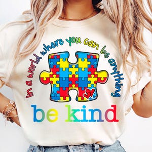 Pu&ograve; includere: Una maglietta bianca con un design a pezzo di puzzle colorato e il testo "in a world where you can be anything be kind".