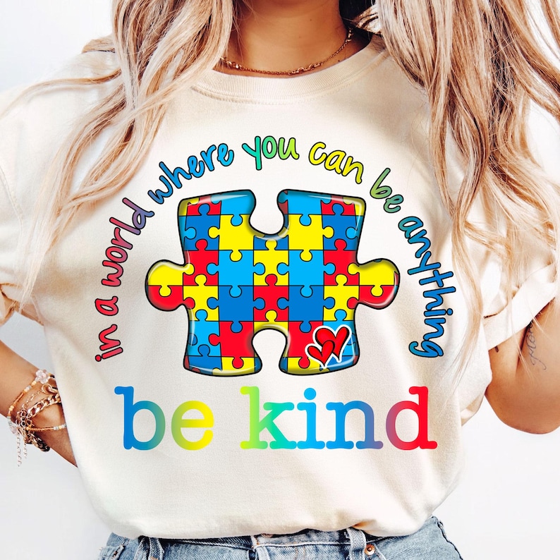 Pu&ograve; includere: Una maglietta bianca con un design a pezzo di puzzle colorato e il testo "in a world where you can be anything be kind".