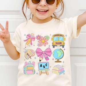 May include: Cream-coloured t-shirt with a colourful school-themed graphic. The design includes a pencil, flowers, school bus, paint palette, bow, globe, backpack, ABC chalkboard, and books. The shirt is worn by a young child.