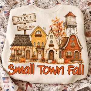 May include: A cream-colored sweatshirt featuring a graphic of a small town in autumn with houses, a water tower, and pumpkins. A wooden sign displays the text 37203 and Nashville, TN. The text Small Town Fall is printed in orange at the bottom.