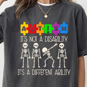 Puede incluir: Camiseta gris con un diseño de pieza de rompecabezas arcoíris que dice "AUTISMO" y el texto "No es una discapacidad, es una habilidad diferente". La camiseta también presenta cuatro figuras de esqueleto, una de las cuales está haciendo una pose de dab.