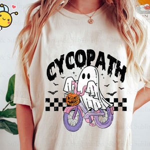 May include: A white t-shirt with a black and white graphic of a ghost riding a bicycle with a pumpkin basket. The text "CYCOPATH" is printed in black above the graphic.
