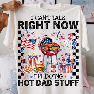 May include: White t-shirt with a graphic design featuring a barbecue scene with an American flag, food, and the text "I CAN'T TALK RIGHT NOW I'M DOING HOT DAD STUFF". The design includes a grill, burgers, hot dogs, and popcorn.