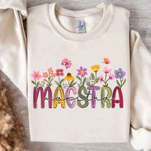 Puede incluir: Una sudadera de color crema con la palabra "MAESTRA" en letras de colores con un diseño floral debajo. Las flores son rosas, moradas, amarillas y verdes.