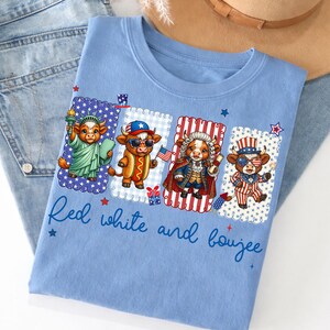 May include: Light blue t-shirt featuring a patriotic design with four cartoon cows dressed in American-themed outfits. The shirt has the text "Red white and boujee" in a script font. The shirt is folded on top of a pair of blue jeans.