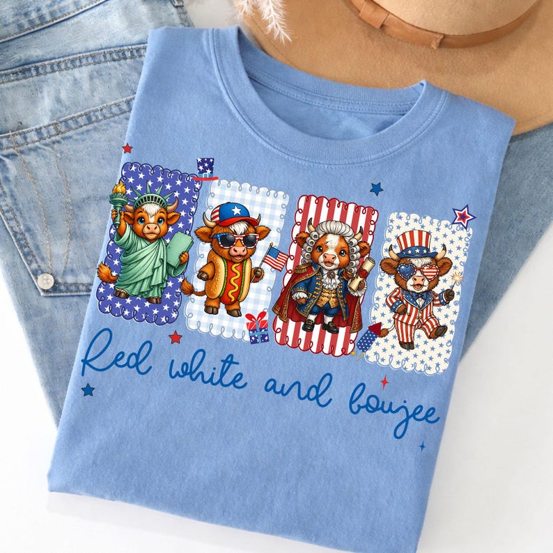 May include: Light blue t-shirt featuring a patriotic design with four cartoon cows dressed in American-themed outfits. The shirt has the text "Red white and boujee" in a script font. The shirt is folded on top of a pair of blue jeans.