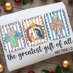 May include: White towel featuring three framed illustrations with a religious theme. The illustrations depict an angel, a nativity scene, and a cross with a bow. The text "the greatest gift of all" and "MATTHEW 1:21" are printed on the towel.