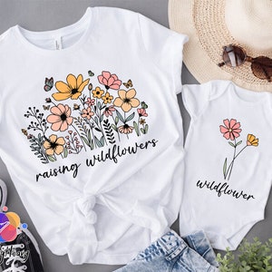 May include: White t-shirts with floral designs and the text "raising wildflowers" and "wildflower".