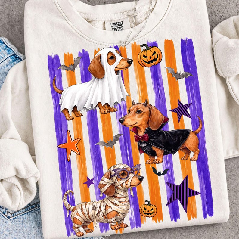 May include: A cream-colored crewneck sweatshirt featuring a graphic of three dachshunds in Halloween costumes: one as a ghost, one as a vampire with a cape, and one as a mummy. The background has vertical orange and purple brushstroke stripes.