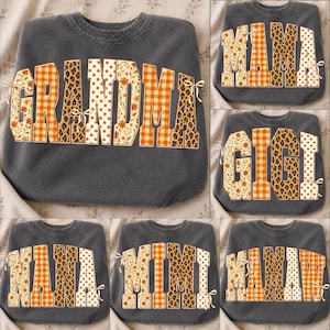 May include: A charcoal gray crewneck sweatshirt featuring large block letters that spell out various names like GRANDMA, MAMA, GIGI, NANA, MIMI, and MAMAW. The letters have a mixed pattern of orange plaid, leopard print, polka dots, and floral.