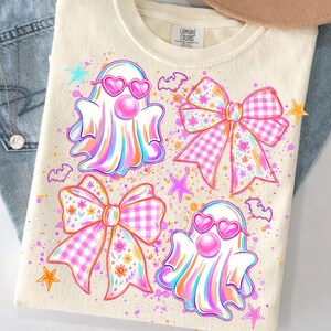 May include: A cream-colored t-shirt with a graphic print featuring two ghosts wearing pink heart-shaped sunglasses and blowing bubblegum, two large bows with pink gingham and floral patterns, small purple bats, and orange and pink stars.