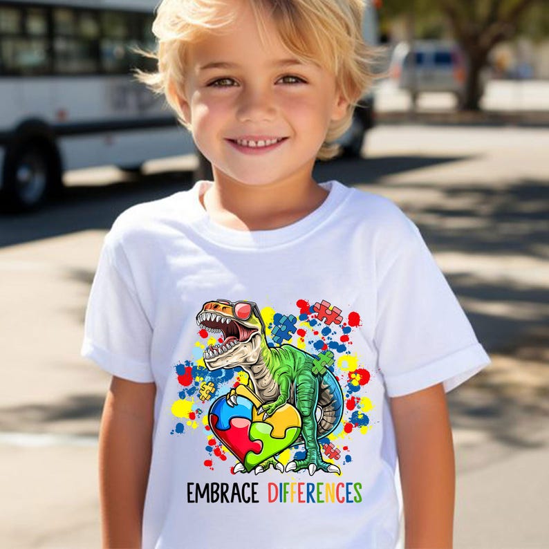 Pu&ograve; includere: Maglietta bianca con una grafica colorata di un dinosauro con occhiali da sole che tiene un cuore a forma di pezzo di puzzle. Il testo "EMBRACE DIFFERENCES" &egrave; stampato sotto la grafica.