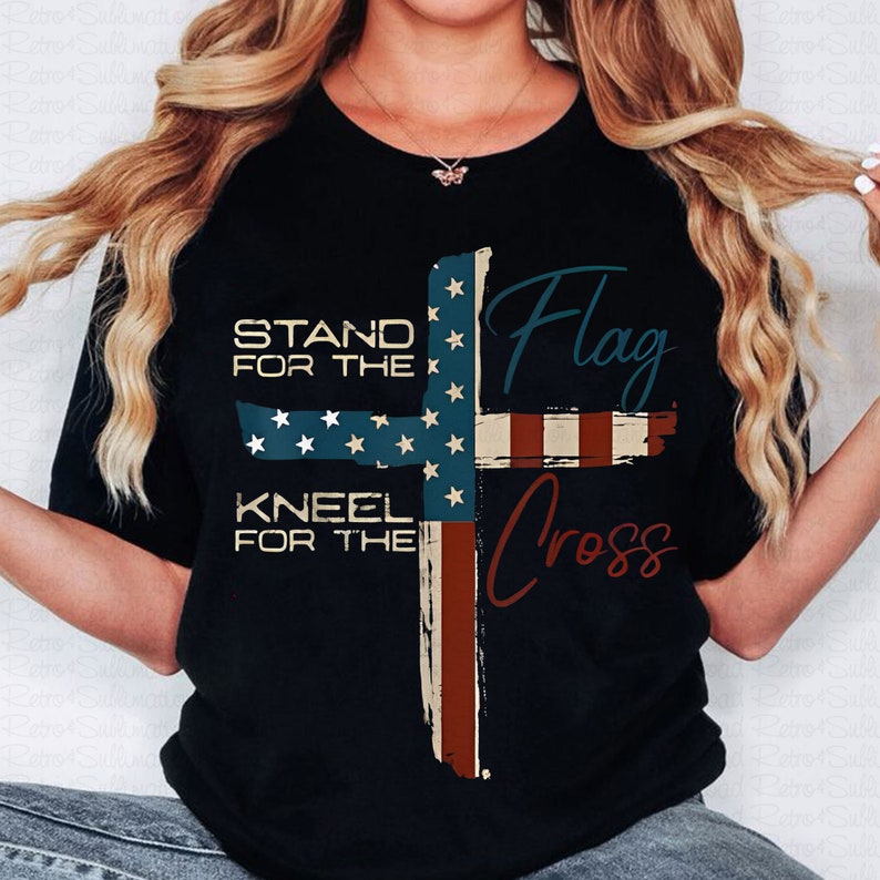 May include: Black t-shirt with a graphic of an American flag in the shape of a cross. The text "Stand for the Flag, Kneel for the Cross" is printed on the shirt.