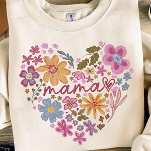 Könnte beinhalten: Ein weißer Pullover mit einem floralen Herzdesign und dem Wort "mama" in rosa Schrift. Das Herz besteht aus verschiedenen Blumen in den Farben Rosa, Gelb, Blau und Grün.