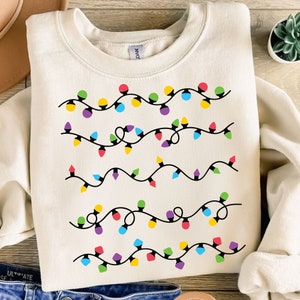 Puede incluir: Sudadera de color crema con un diseño de luces de colores. El diseño incluye múltiples hilos de luces en varios colores, incluyendo rojo, amarillo, verde, morado y azul. La sudadera está hecha de un material suave y es adecuada para uso casual.