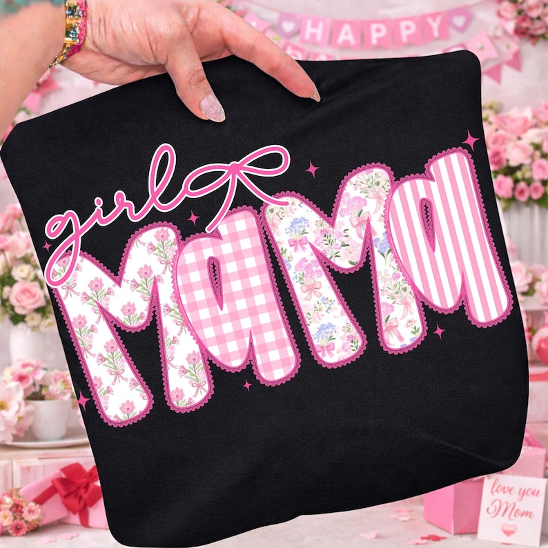 Puede incluir: Camiseta negra con las palabras "girl mama" en letras rosas. Las letras est&aacute;n decoradas con estampados florales, cuadros rosas y rayas rosas. Un lazo rosa est&aacute; encima de la palabra "mama".