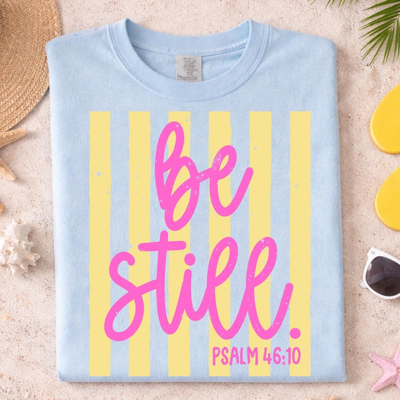 May include: Light blue t-shirt with yellow vertical stripes and the words "be still" in pink script. The text "PSALM 46:10" is below the words. The shirt is displayed on a sandy surface with beach accessories.