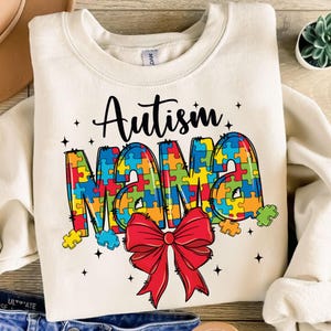 May include: A white sweatshirt with the text "Autism Mama" in black. The word "Mama" is made up of colorful puzzle pieces and a red bow.