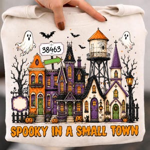 Peut inclure: Un sweat-shirt crème avec une illustration de ville effrayante aux bâtiments orange et violets, un château d'eau, des fantômes, des chauves-souris et des arbres nus. Un panneau indique 38463 avec une flèche verte. Le texte indique SPOOKY IN A SMALL TOWN.