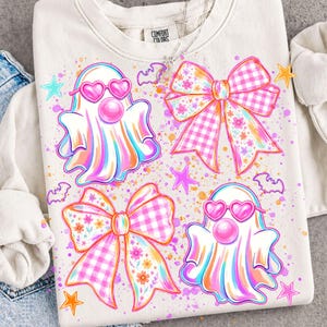 May include: A cream-colored crewneck sweatshirt featuring a graphic print of two ghosts wearing pink heart-shaped glasses and two large bows with pink gingham and floral patterns, accented by small stars, bats, and colorful paint splatters.